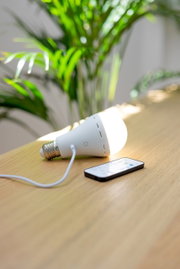Akku-Leuchtmittel auf einem Tisch liegend, mit angeschlossenem Ladekabel und Fernbedienung, seitliche Perspektive