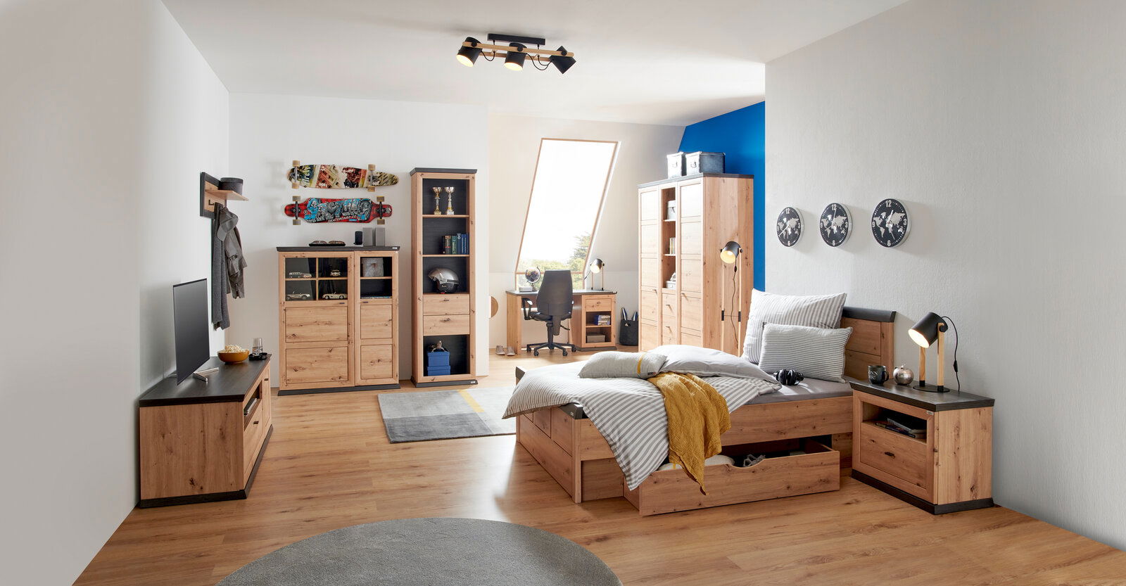 Modernes Schlafzimmer mit Holzmöbeln, darunter ein Bett mit Stauraum, ein Schreibtisch mit Stuhl und ein Kleiderschrank, aus der Perspektive von der Zimmerecke aus gesehen.