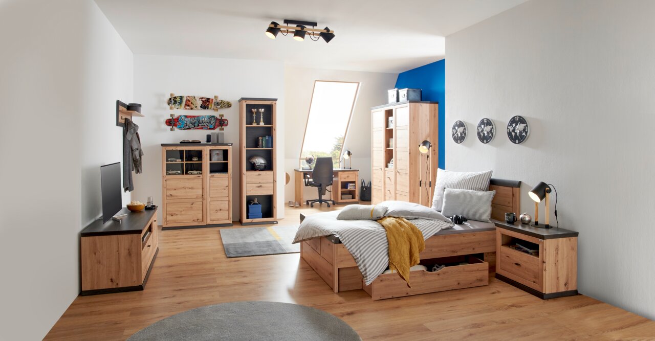 Hom´in Plank BENTON Modernes Schlafzimmer mit Holzmöbeln, darunter ein Bett mit Stauraum, ein Schreibtisch mit Stuhl und ein Kleiderschrank, aus der Perspektive von der Zimmerecke aus gesehen.