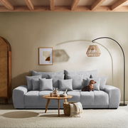 Großes, graues Big-Sofa in einem modernen Wohnzimmer, frontal fotografiert. Dekoriert mit mehreren Kissen, einem kleinen Holztisch mit Karaffe und Vase, sowie einem Hund, der auf dem Sofa sitzt. Im Hintergrund eine Stehlampe und ein gerahmtes Bild an der