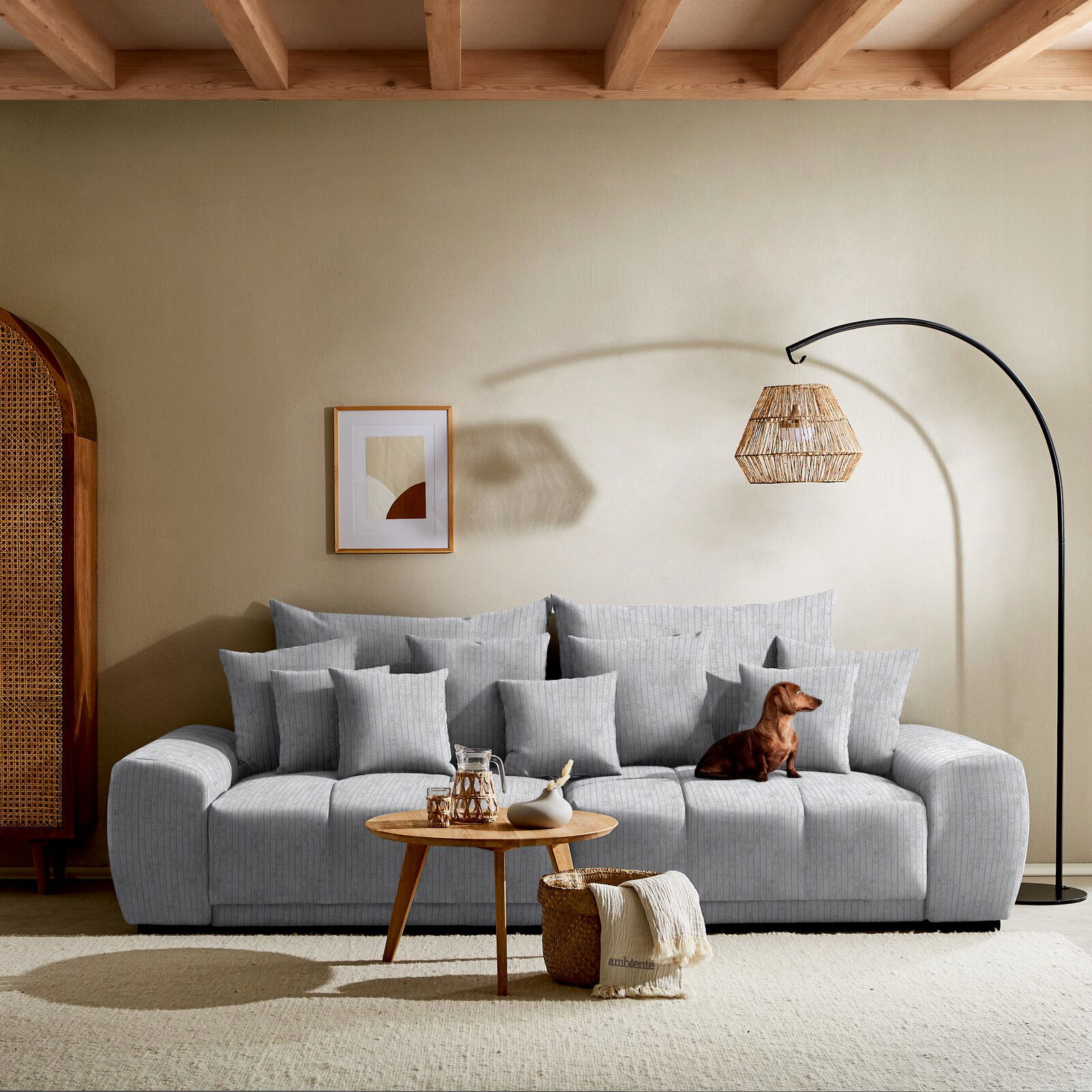Großes, graues Big-Sofa in einem modernen Wohnzimmer, frontal fotografiert. Dekoriert mit mehreren Kissen, einem kleinen Holztisch mit Karaffe und Vase, sowie einem Hund, der auf dem Sofa sitzt. Im Hintergrund eine Stehlampe und ein gerahmtes Bild an der