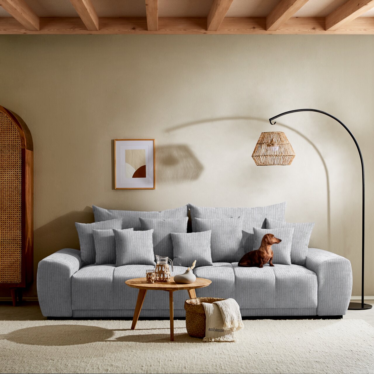 Großes, graues Big-Sofa in einem modernen Wohnzimmer, frontal fotografiert. Dekoriert mit mehreren Kissen, einem kleinen Holztisch mit Karaffe und Vase, sowie einem Hund, der auf dem Sofa sitzt. Im Hintergrund eine Stehlampe und ein gerahmtes Bild an der 