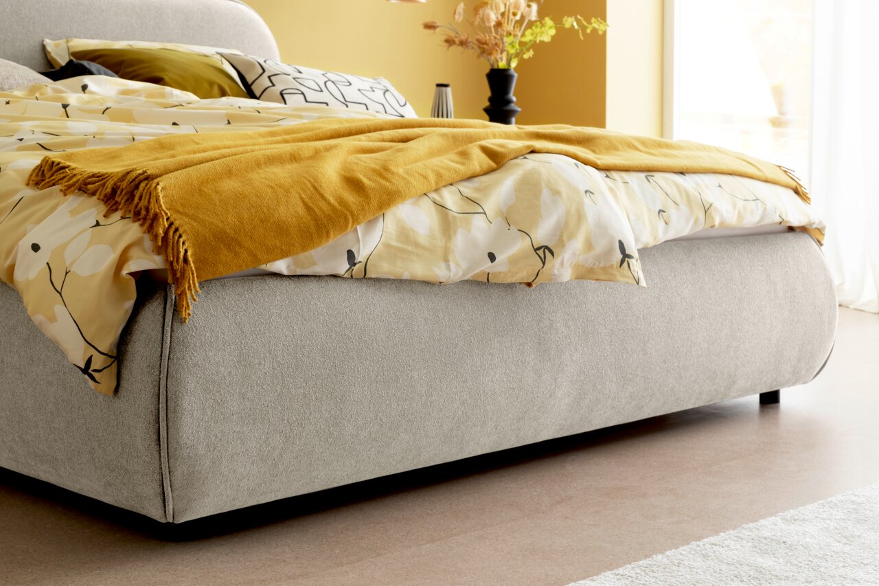 Seitliche Perspektive auf ein graues Polsterbettgestell LISSABON mit gelber Decke und gemusterten Kissen.