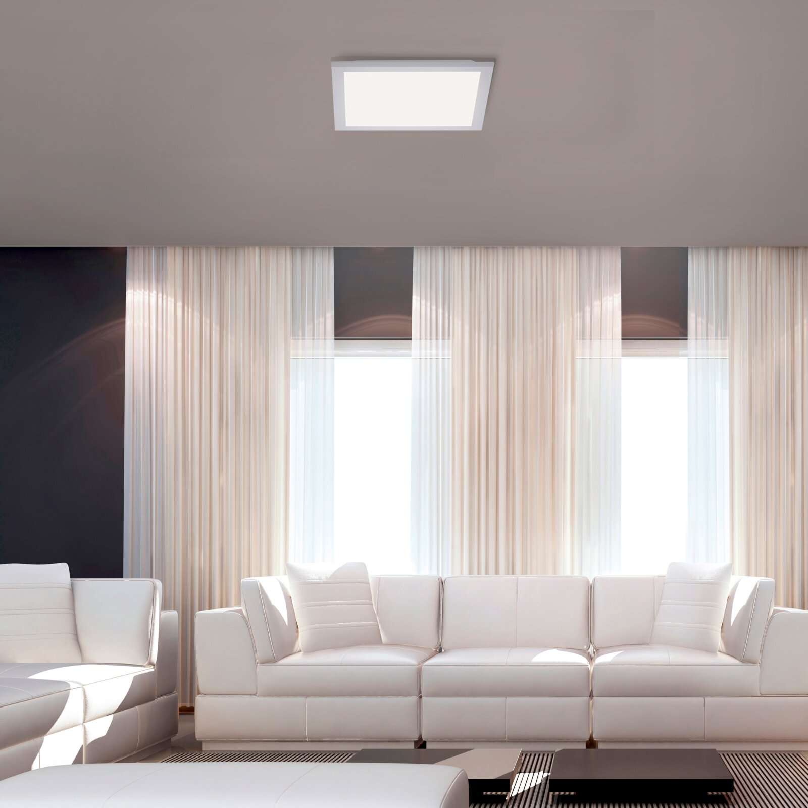 Quadratische LED-Deckenleuchte in Weiß an der Zimmerdecke über einem modernen Wohnzimmer mit weißen Sofas und großen Fenstern, Ansicht von unten/leicht frontal zur Decke.