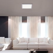 Quadratische LED-Deckenleuchte in Weiß an der Zimmerdecke über einem modernen Wohnzimmer mit weißen Sofas und großen Fenstern, Ansicht von unten/leicht frontal zur Decke.