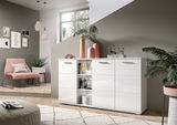 Weiße Kommode mit glänzender Oberfläche, dekoriert mit Vasen und Büchern, in einem modernen Wohnzimmer mit Pflanzen und Kunstwerken an der Wand. Perspektive von vorne.