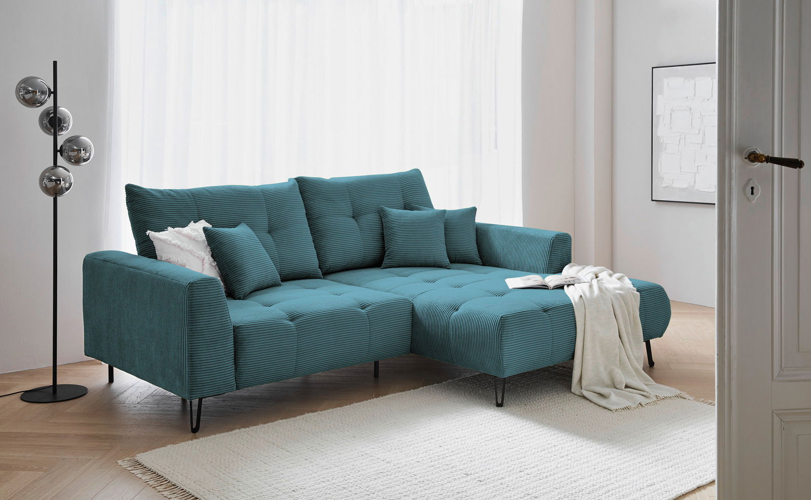 Moderne, türkisfarbene Eckcouch mit Ottomane rechts, dekoriert mit Kissen und Decke, in einem hellen Wohnzimmer mit Holzfußboden, seitliche Perspektive.