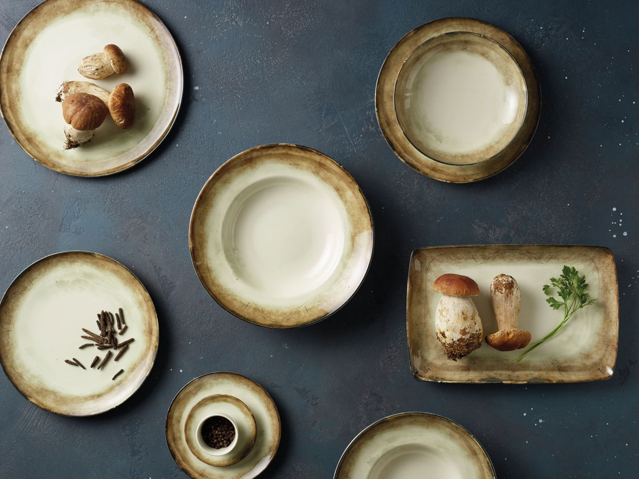 Vogelperspektive auf ein 12-teiliges Tellerset 'Shade Earth' mit erdigen Farbtönen, dekoriert mit Pilzen und Gewürzen.