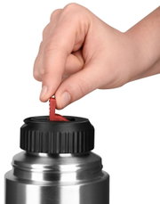Close-up van een hand die de sluiting van een zilveren thermosfles met een zwarte dop en een rode hendel opent.
