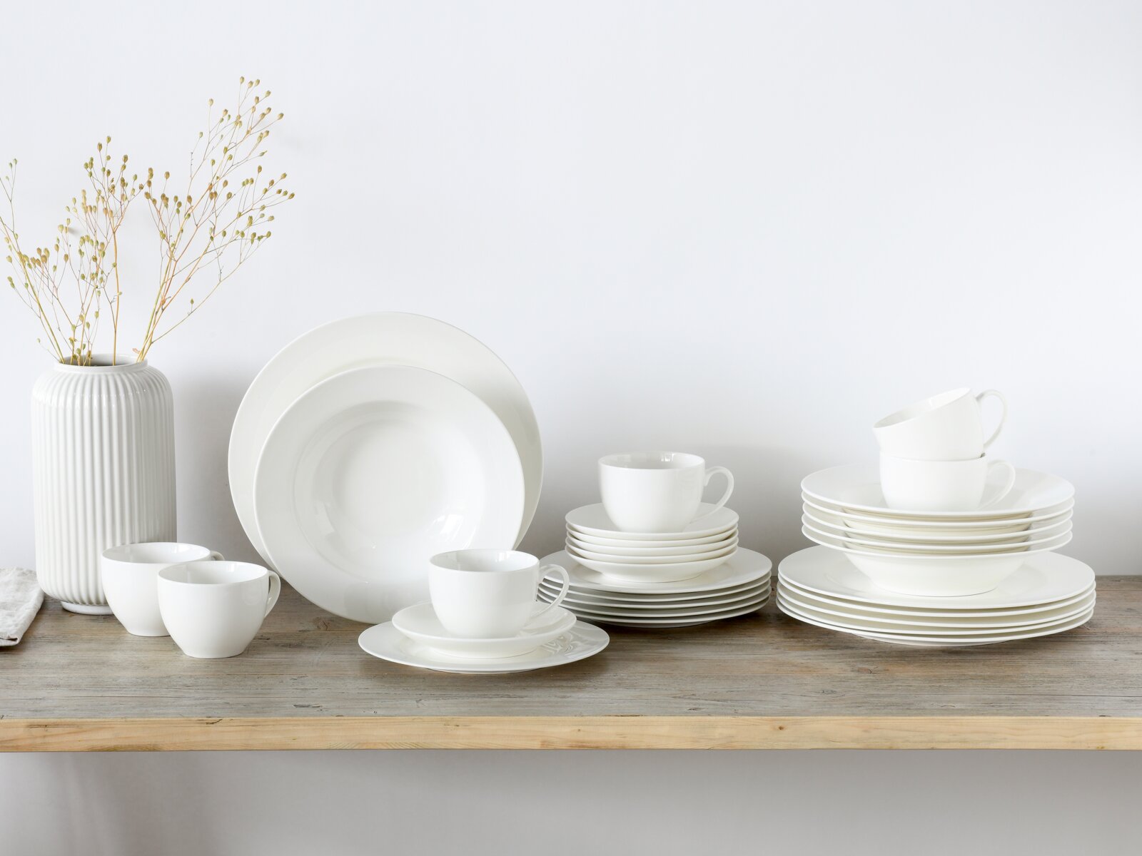 Creatable Kombiservice 30-tlg FLEUR ROYAL Weißes Porzellan-Geschirrset mit Tellern, Schalen und Tassen auf einem Holztisch, seitliche Frontansicht mit leicht erhöhter Perspektive; links eine geriffelte weiße Vase mit Trockenblumen vor hellem Hintergrund.