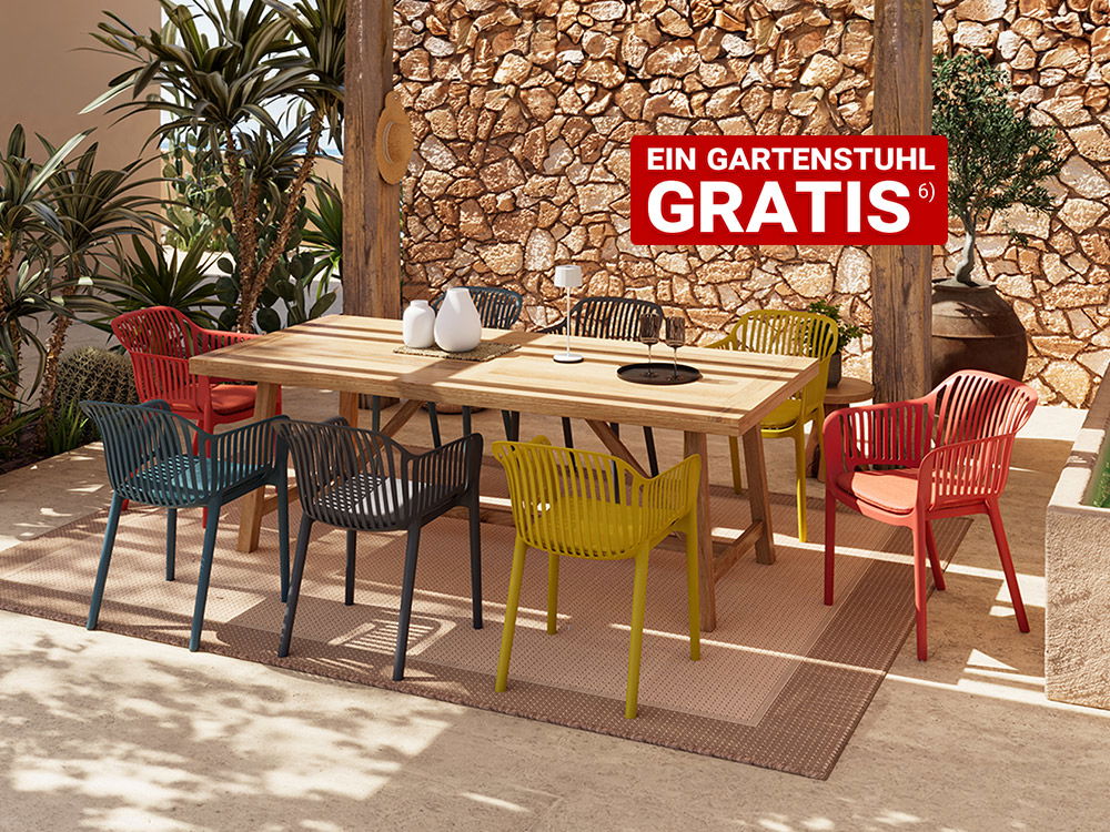 Großer rechteckiger Holztisch mit mehreren bunten Kunststoffstühlen auf einer Terrasse vor einer Steinwand und Pflanzen