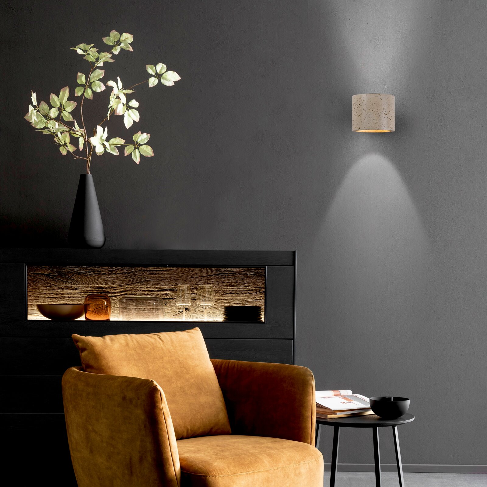 Moderne Wandlampe mit zylindrischem, gesprenkeltem Schirm in Beige/Creme, die warmes Licht nach unten auf eine graue Wand wirft; frontale Ansicht leicht von links.