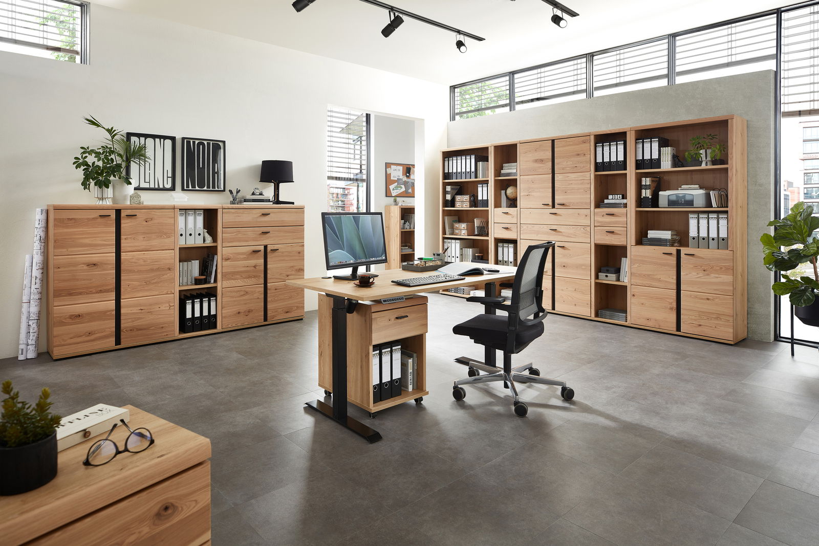 Moderne Büromöbel in einem hellen Raum mit großen Fenstern, bestehend aus einem Schreibtisch mit Rollcontainer, mehreren Aktenschränken und Regalen aus Holz. Perspektive: Frontalansicht.