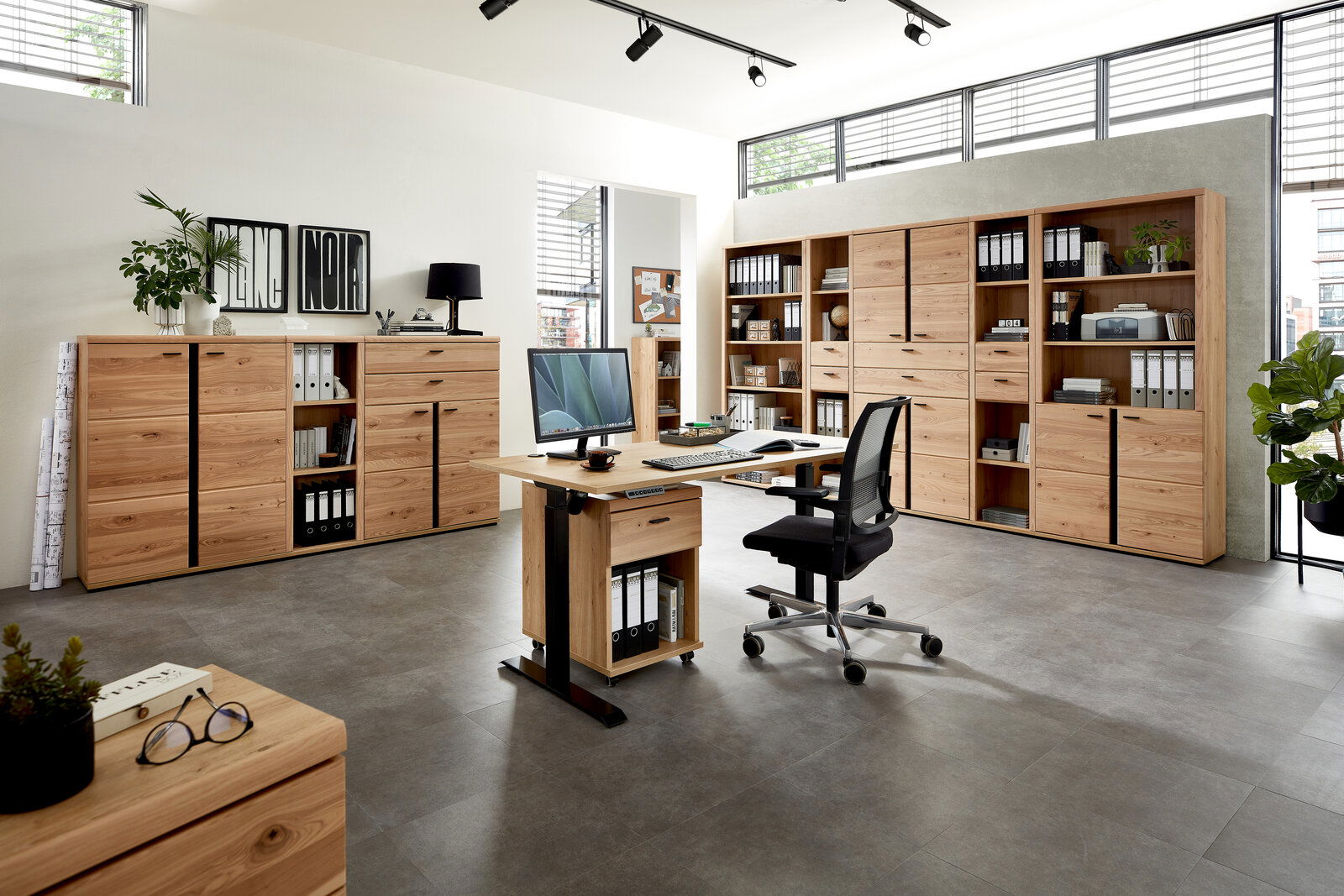 Moderne Büromöbel in einem hellen Raum mit großen Fenstern, bestehend aus einem Schreibtisch mit Rollcontainer, mehreren Aktenschränken und Regalen aus Holz. Perspektive: Frontalansicht.