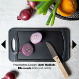 Schwarzes Schneidebrett mit Anti-Rutsch-Elementen, darauf geschnittene rote Zwiebel und kleines Messer, von oben fotografiert.