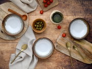 Drei Keramikschalen mit weißer Glasur und braunem Rand auf rustikalem Holztisch, umgeben von goldfarbenen Löffeln, Oliven, Kirschtomaten und einem Glas – Draufsicht (Flatlay)