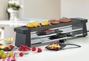 Raclette-Grill mit Alugrillplatte, seitliche Perspektive, mit verschiedenen Speisen wie Garnelen, Würstchen und Gemüse auf der Grillfläche und in Pfännchen darunter.