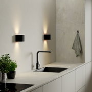 Moderne schwarze Wandleuchten in rechteckiger Form mit Up-&-Down-Light über einer Küchenarbeitsplatte mit schwarzer Armatur und Spüle, Perspektive: leicht seitliche Frontansicht.