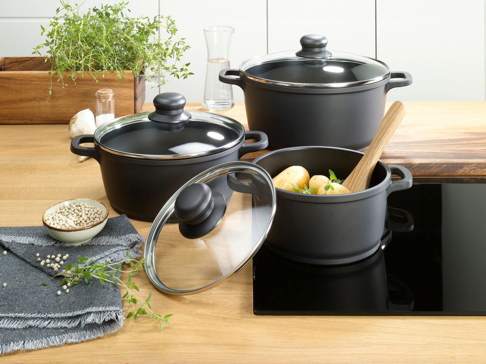 Homeware Topfset 3-tlg. Dreiteiliges Topfset in moderner Küche, bestehend aus schwarzen Töpfen mit Glasdeckeln, gefüllt mit Kartoffeln und Kräutern, aus der Vogelperspektive fotografiert.