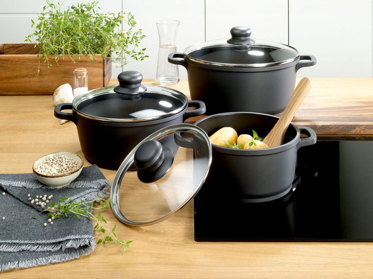 Dreiteiliges Topfset in moderner Küche, bestehend aus schwarzen Töpfen mit Glasdeckeln, gefüllt mit Kartoffeln und Kräutern, aus der Vogelperspektive fotografiert.
