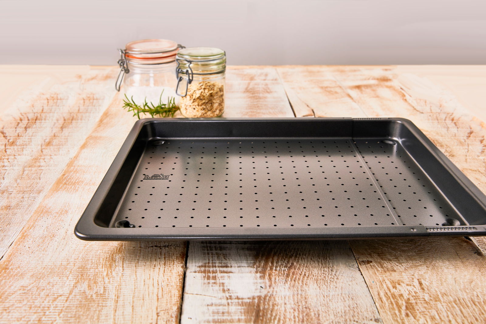 Perforiertes Backblech auf einem Holztisch, seitliche Perspektive, mit Gewürzgläsern im Hintergrund.