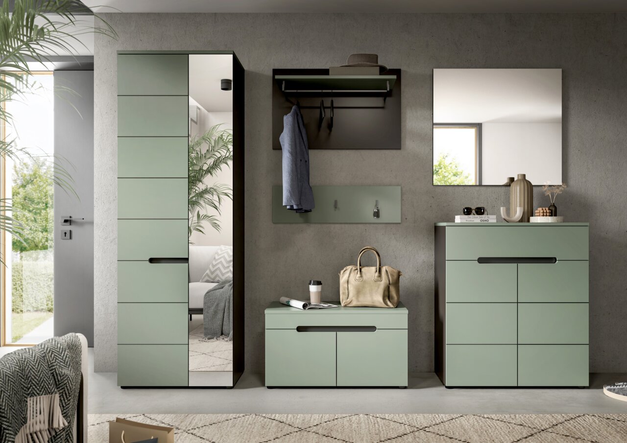 Frontalansicht eines modernen Flurmöbel-Sets in Salbeigrün mit hohem Garderobenschrank mit Spiegel, Wandgarderobe mit Hutablage und Haken, Sitzbank sowie Kommode unter einem Wandspiegel vor grauer Betonwand.