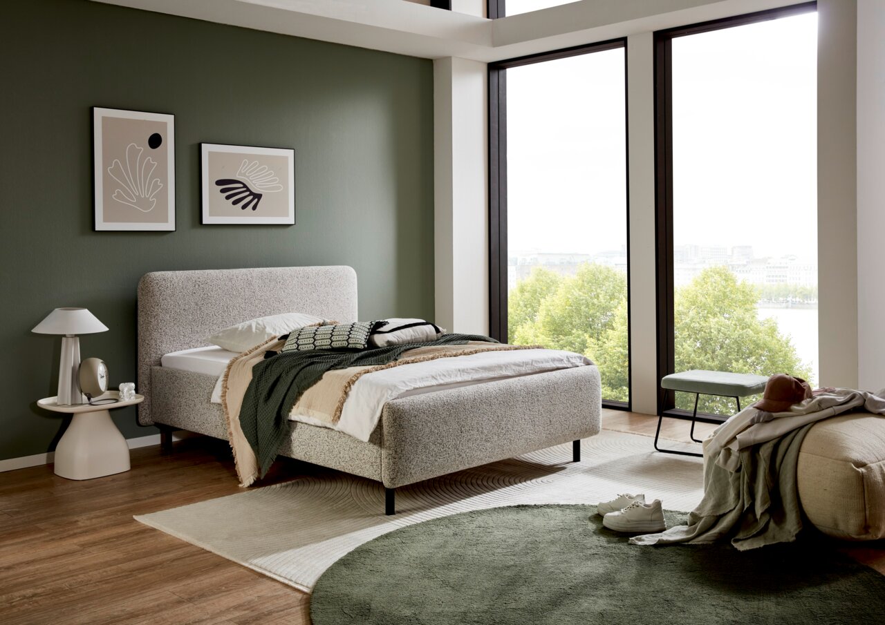 Modernes Polsterbett in einem stilvollen Schlafzimmer mit grüner Wand, großen Fenstern und minimalistischer Dekoration, seitliche Perspektive.