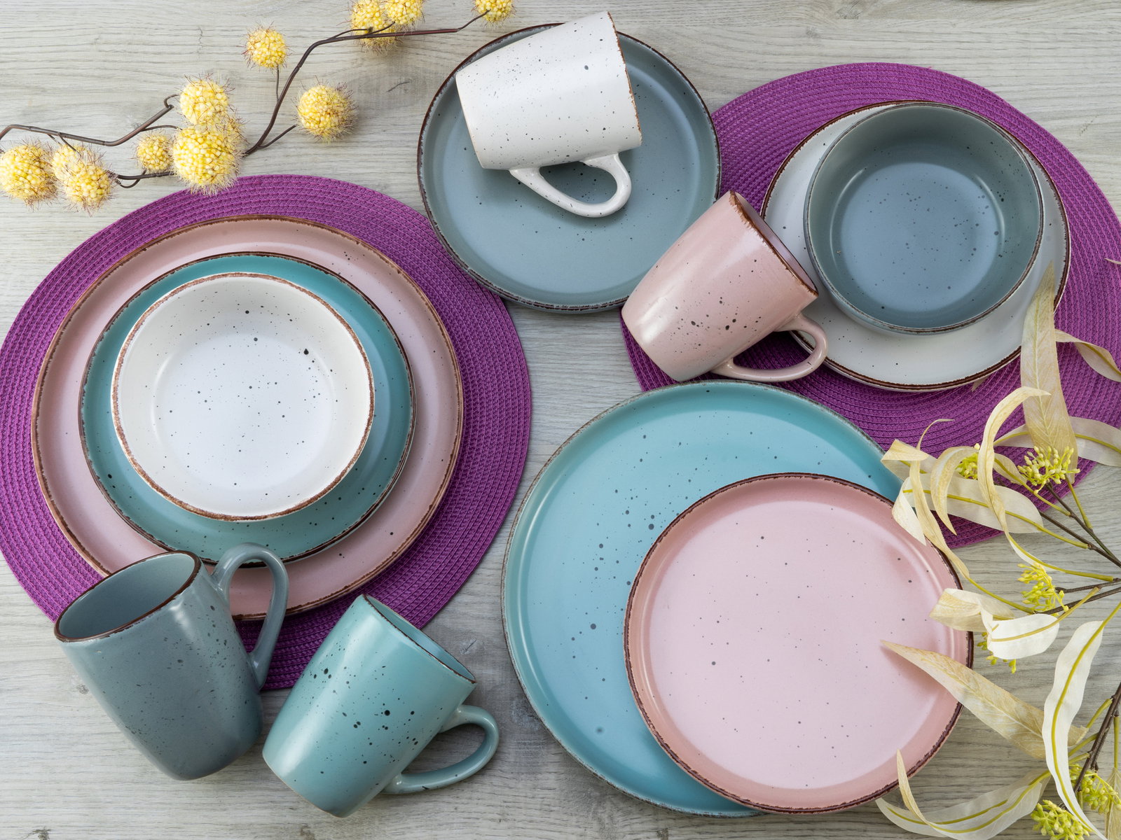 Vogelperspektive auf ein 16-teiliges Kombiservice in modernen Pastellfarben, bestehend aus Tellern, Schalen und Tassen, arrangiert auf einem Holztisch mit lila Platzsets und gelben Blumen.