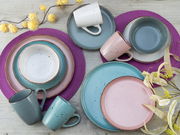 Vogelperspektive auf ein 16-teiliges Kombiservice in modernen Pastellfarben, bestehend aus Tellern, Schalen und Tassen, arrangiert auf einem Holztisch mit lila Platzsets und gelben Blumen.
