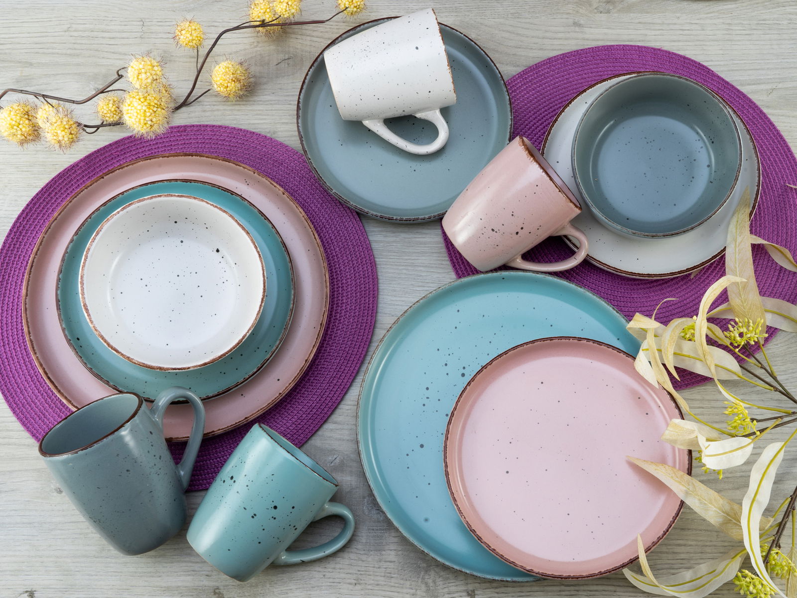 Vogelperspektive auf ein 16-teiliges Kombiservice in modernen Pastellfarben, bestehend aus Tellern, Schalen und Tassen, arrangiert auf einem Holztisch mit lila Platzsets und gelben Blumen.