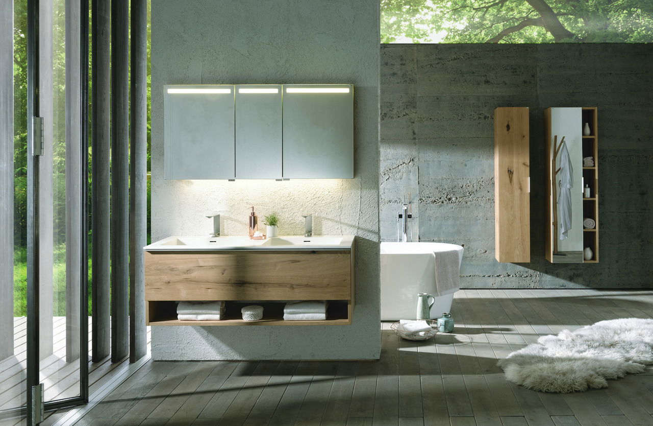 Moderne Badkombination mit hängendem Waschtisch aus Holz, beleuchtetem Spiegel und freistehender Badewanne in einem lichtdurchfluteten Raum mit Blick auf den Garten.
