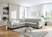 Hellgraues L-förmiges Ecksofa mit gepolsterten Armlehnen in einem hellen Wohnzimmer mit Couchtisch und Teppich, Perspektive: frontale Weitwinkelansicht leicht von links.