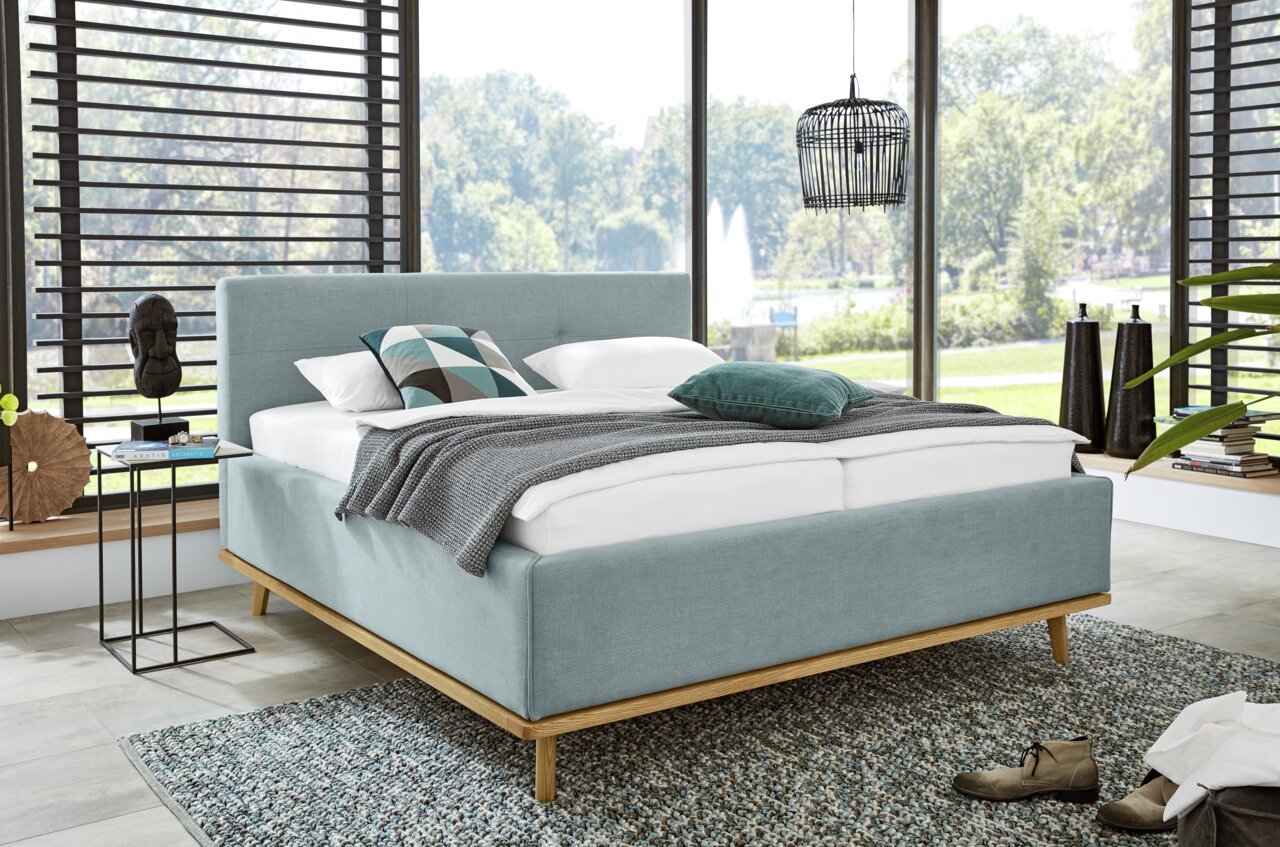 Modern gestoffeerd bed van lichtblauwe stof met houten frame, versierd met kussens en een deken, in een lichte kamer met grote ramen, zijaanzicht.