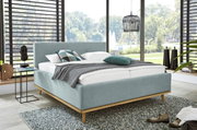 Modern gestoffeerd bed van lichtblauwe stof met houten frame, versierd met kussens en een deken, in een lichte kamer met grote ramen, zijaanzicht.