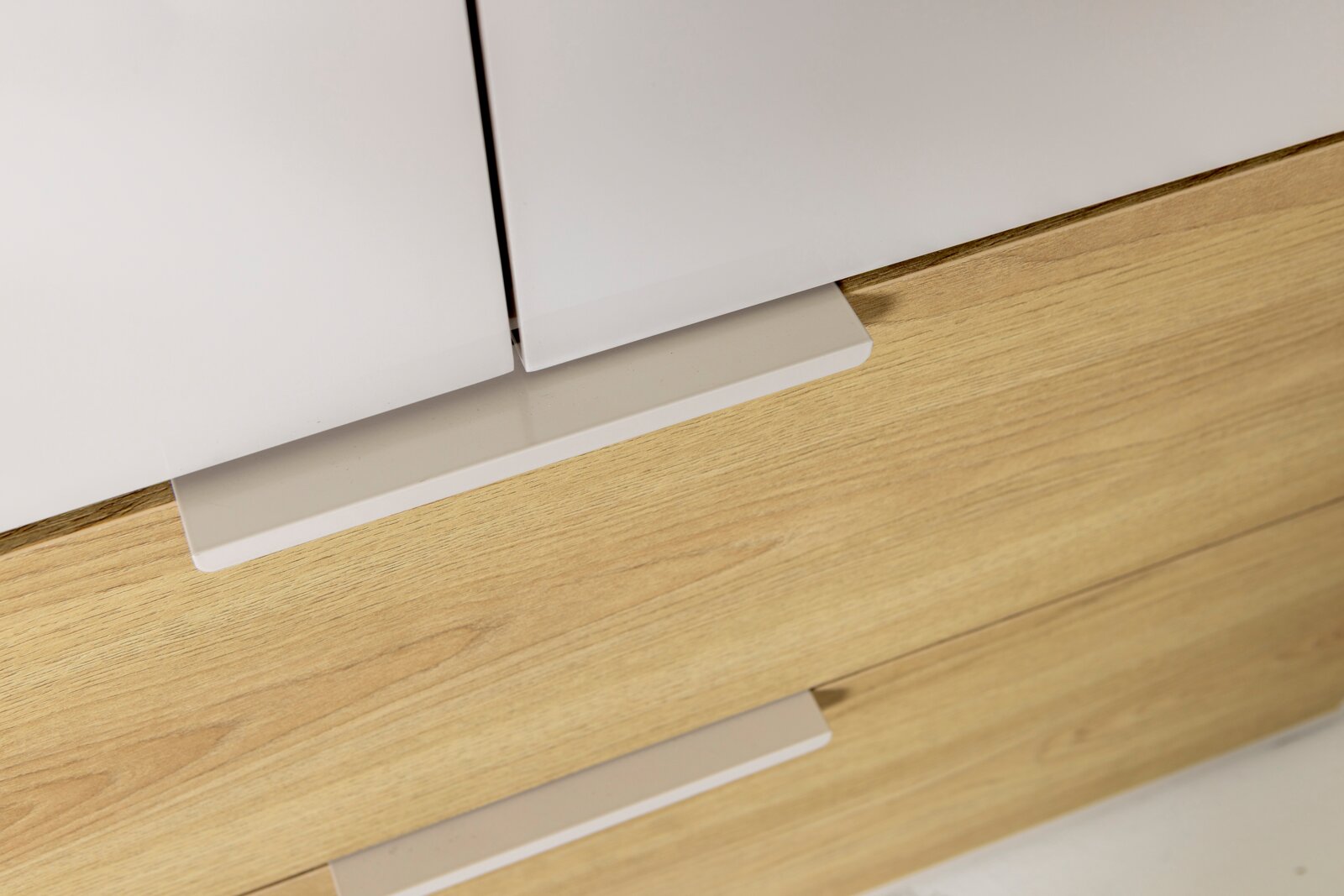 Detailaufnahme aus leicht schräger Frontperspektive: weißes Sideboard/Schrank mit Holzfront und flachen weißen Griffleisten auf hellem Holzboden