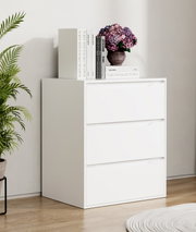 Weiße Kommode mit drei Schubladen in modernem, minimalistischem Design, auf der Ablage Bücher und eine Vase mit lila Hortensien; Aufnahme in leichter Seitenansicht von vorn (3/4-Perspektive) in einem hellen Wohnraum.