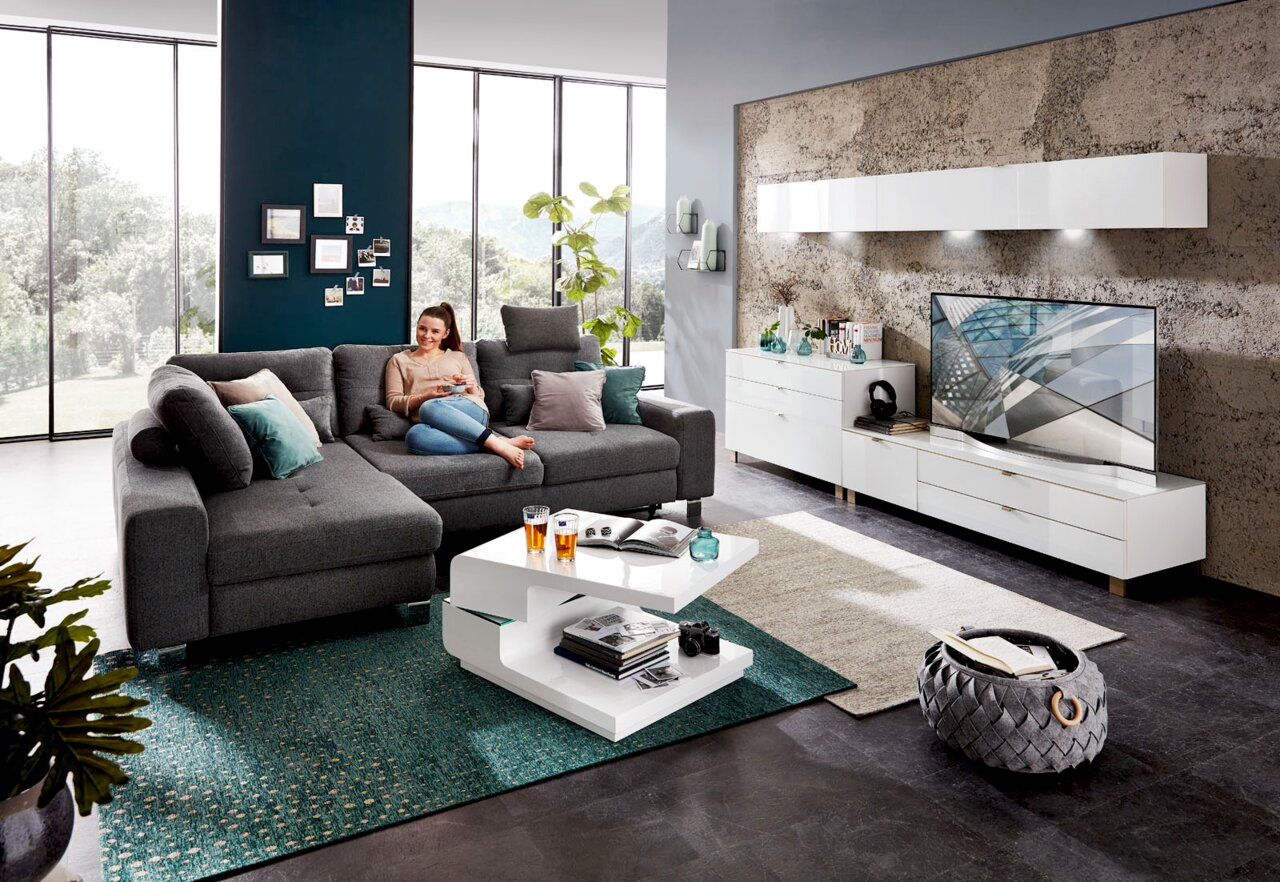 Moderne Wohnzimmeransicht mit grauem Ecksofa, weißem Funktions-Couchtisch und stilvollem Hängeschrank an der Wand.