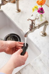 Eine Person wäscht einen schwarzen Mörserstößel unter fließendem Wasser in einem Waschbecken. Der Mörser ist im Hintergrund sichtbar. Perspektive von oben.