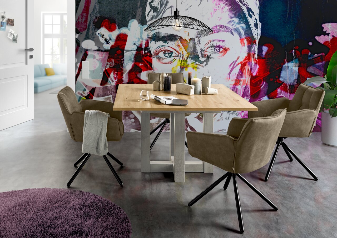 Esszimmer mit einem Holztisch und vier drehbaren Armlehnstühlen der Serie FRISCO von Set One by Musterring, vor einer bunten Wand mit Kunstmotiv. Perspektive: seitlich.