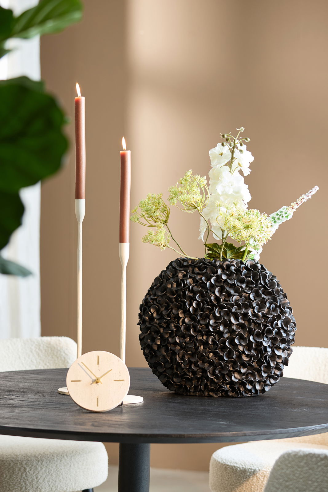 Dekorative Vase mit strukturierter Oberfläche, gefüllt mit Blumen, auf einem Tisch neben zwei brennenden Kerzen und einer kleinen Uhr. Perspektive: Frontalansicht.