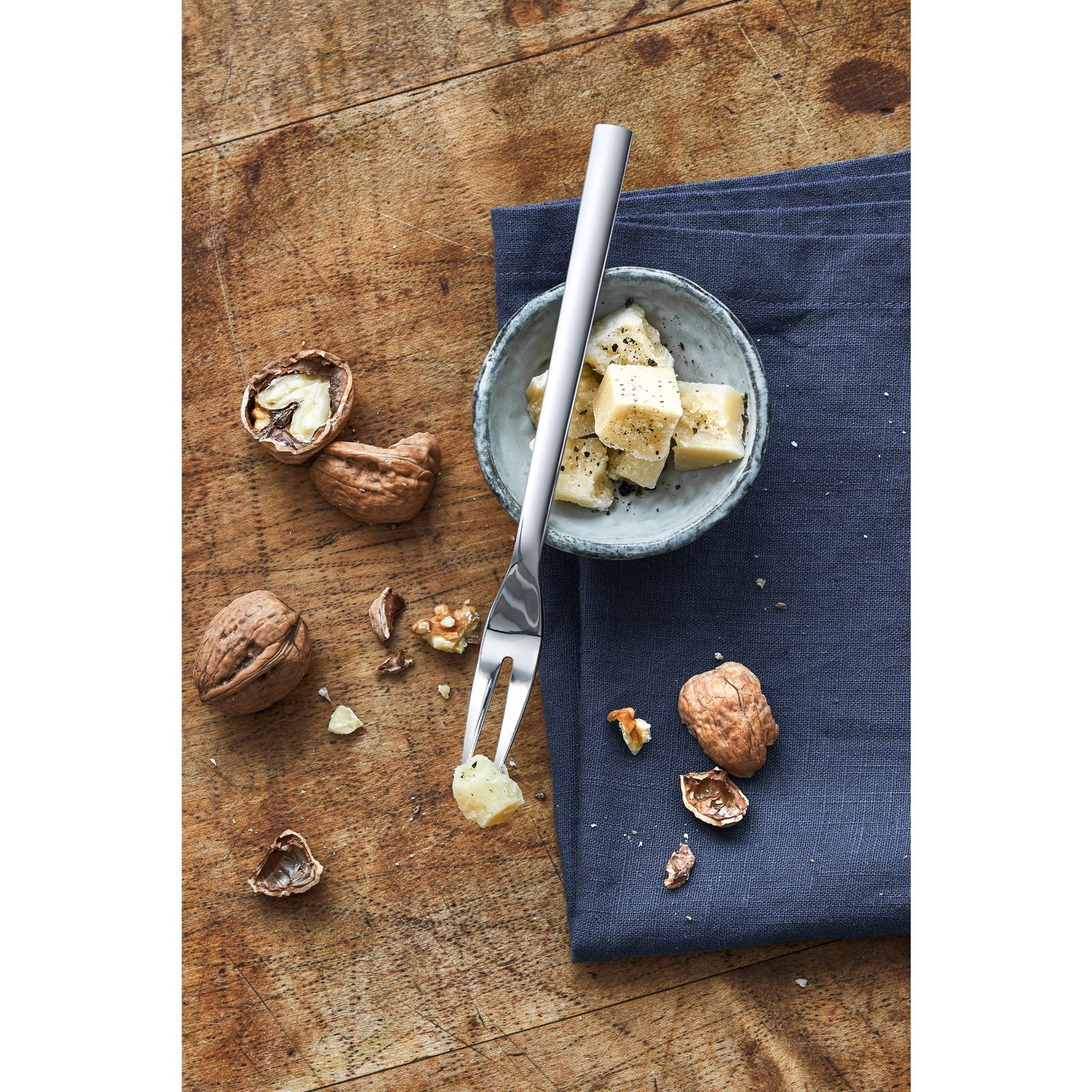 Serviergabel aus Edelstahl auf einem Holztisch, neben einer Schale mit Käsewürfeln und Walnüssen, aus der Vogelperspektive.