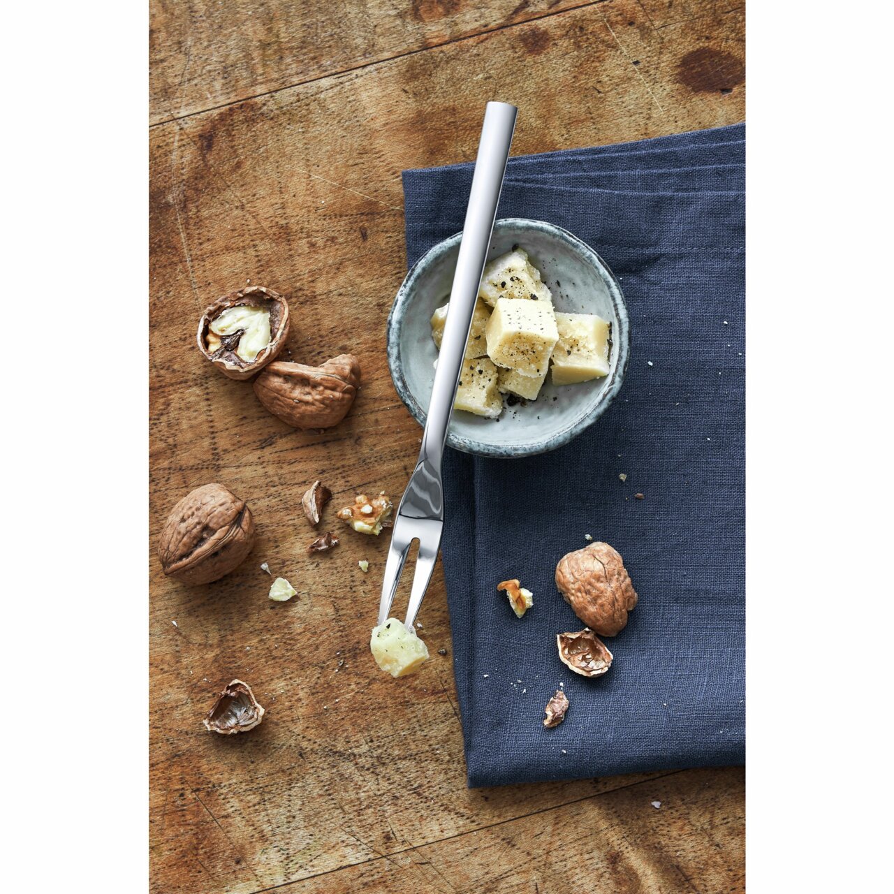 Serviergabel aus Edelstahl auf einem Holztisch, neben einer Schale mit Käsewürfeln und Walnüssen, aus der Vogelperspektive.