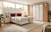 Modernes Schlafzimmer-Set aus gepolstertem Doppelbett, Nachtkommode und großem Drehtürenschrank in warmer Holzoptik mit weißen Hochglanzfronten, aus schräger Vorderansicht.