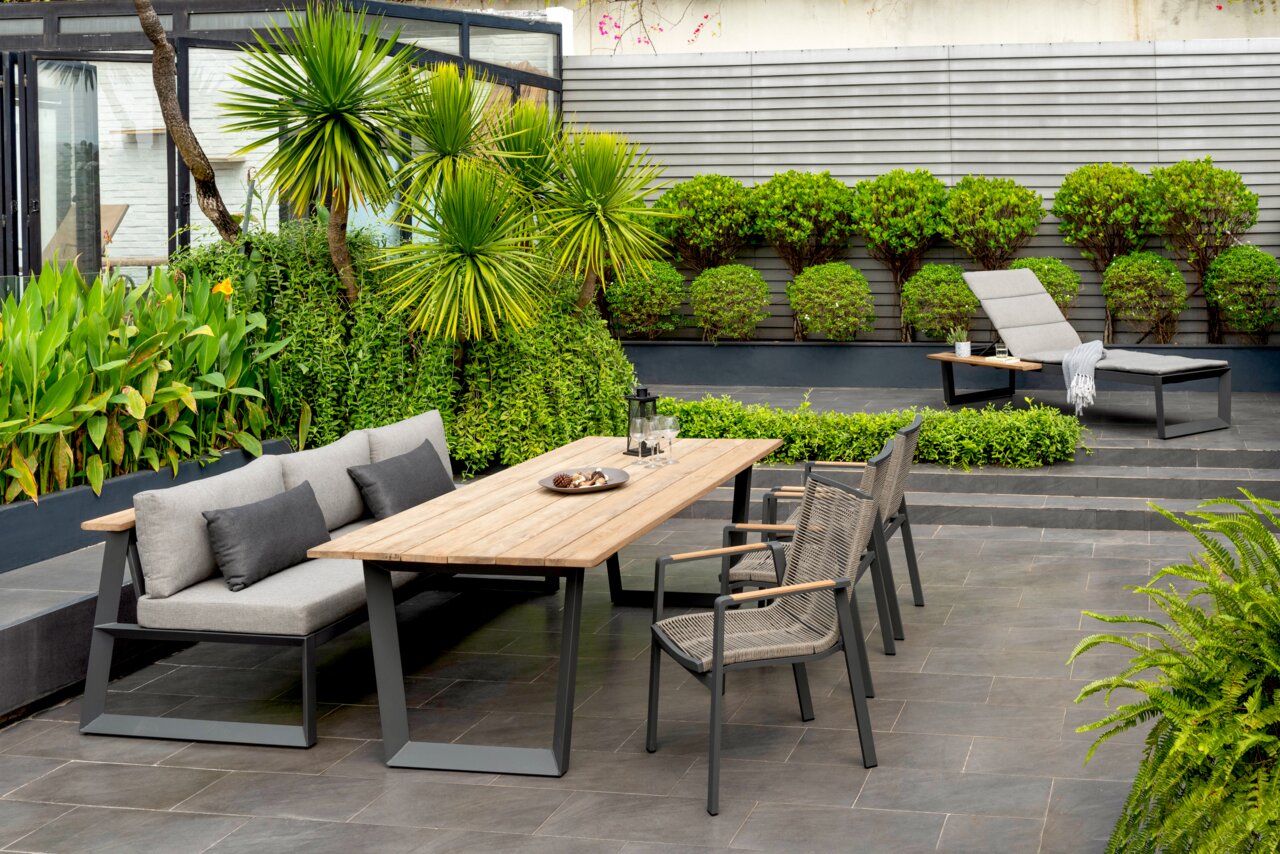 Moderner Gartentisch mit Holzplatte und grauem Metallgestell, umgeben von stilvollen Gartenstühlen und einer Sitzbank, in einem eleganten Garten mit üppiger Bepflanzung, aus der Perspektive von vorne betrachtet.