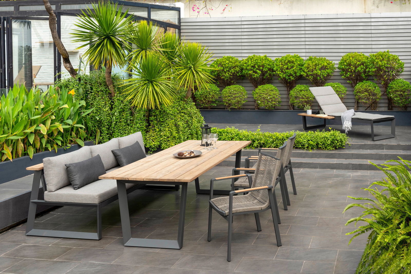 Moderner Gartentisch mit Holzplatte und grauem Metallgestell, umgeben von stilvollen Gartenstühlen und einer Sitzbank, in einem eleganten Garten mit üppiger Bepflanzung, aus der Perspektive von vorne betrachtet.