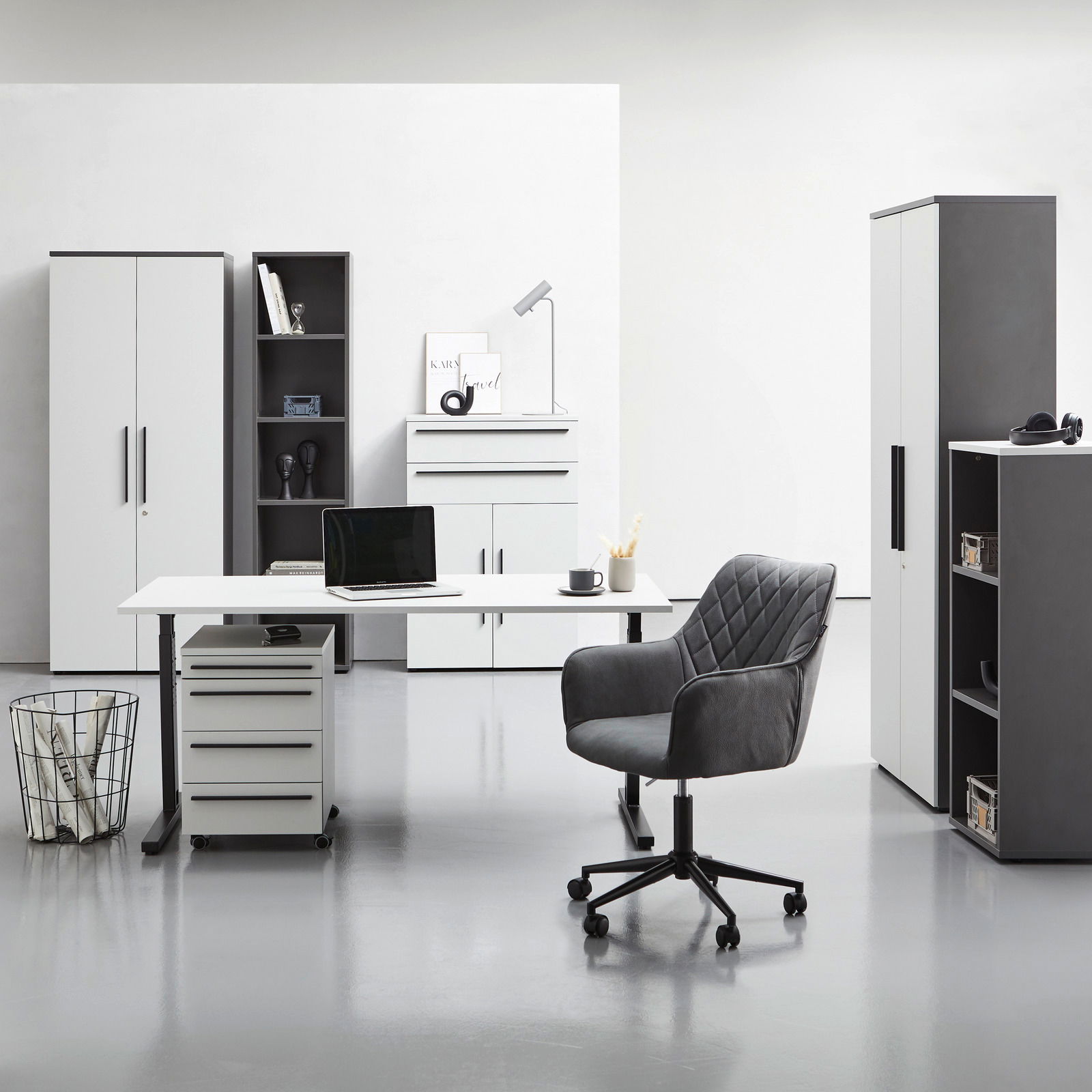 Moderne Büromöbel in einem hellen Raum, bestehend aus einem Winkelschreibtisch mit Rollcontainer, einem Drehstuhl und mehreren Aktenschränken und Regalen, frontal betrachtet.