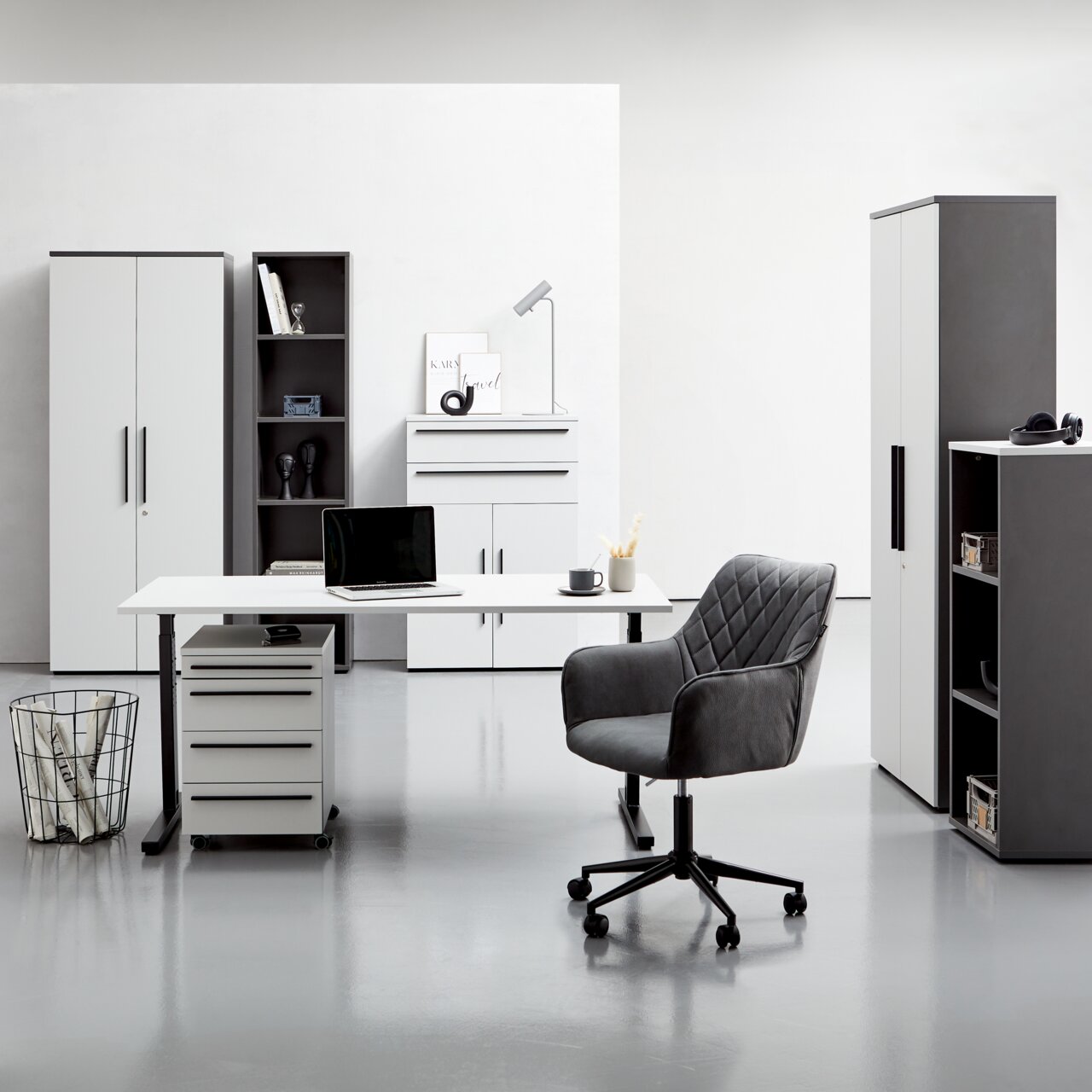 Moderne Büromöbel in einem hellen Raum, bestehend aus einem Winkelschreibtisch mit Rollcontainer, einem Drehstuhl und mehreren Aktenschränken und Regalen, frontal betrachtet.