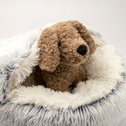 Plüschhund sitzt in einem flauschigen, runden Haustierbett mit weißem und grauem Kunstfell, seitliche Perspektive