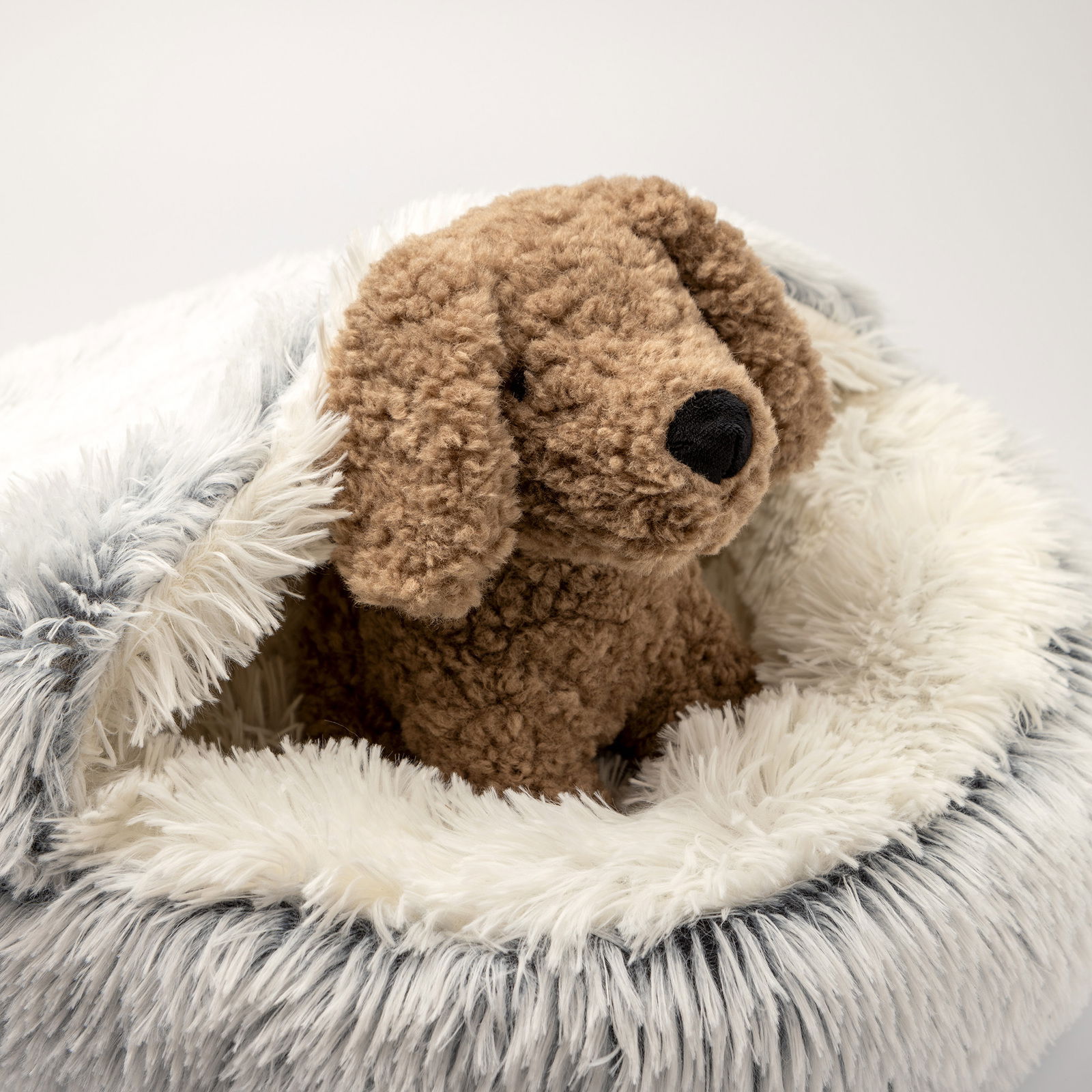 Plüschhund sitzt in einem flauschigen, runden Haustierbett mit weißem und grauem Kunstfell, seitliche Perspektive