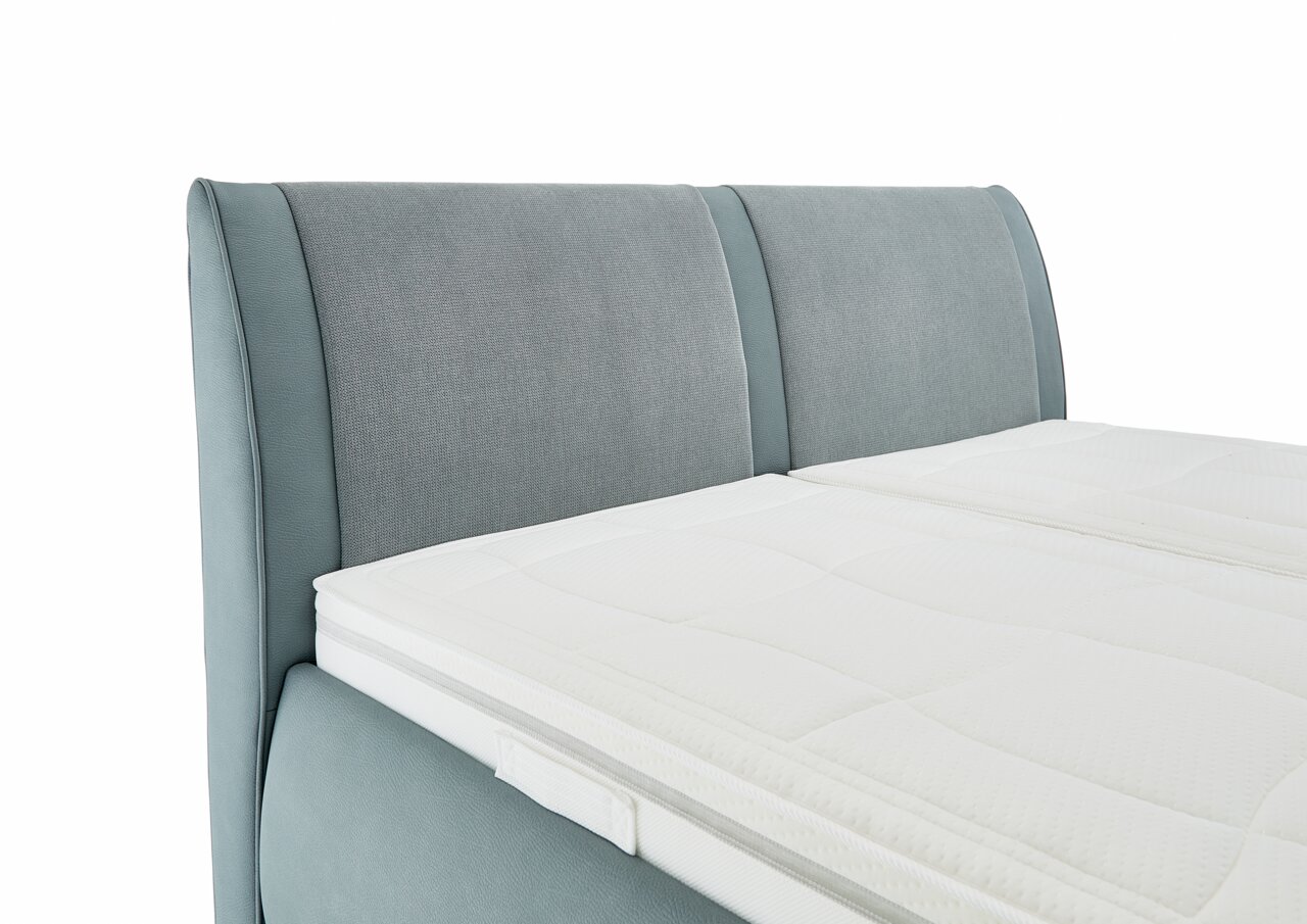 Nahaufnahme eines Polsterbetts mit hellgrauem Kopfteil und weißer Matratze, seitliche Perspektive